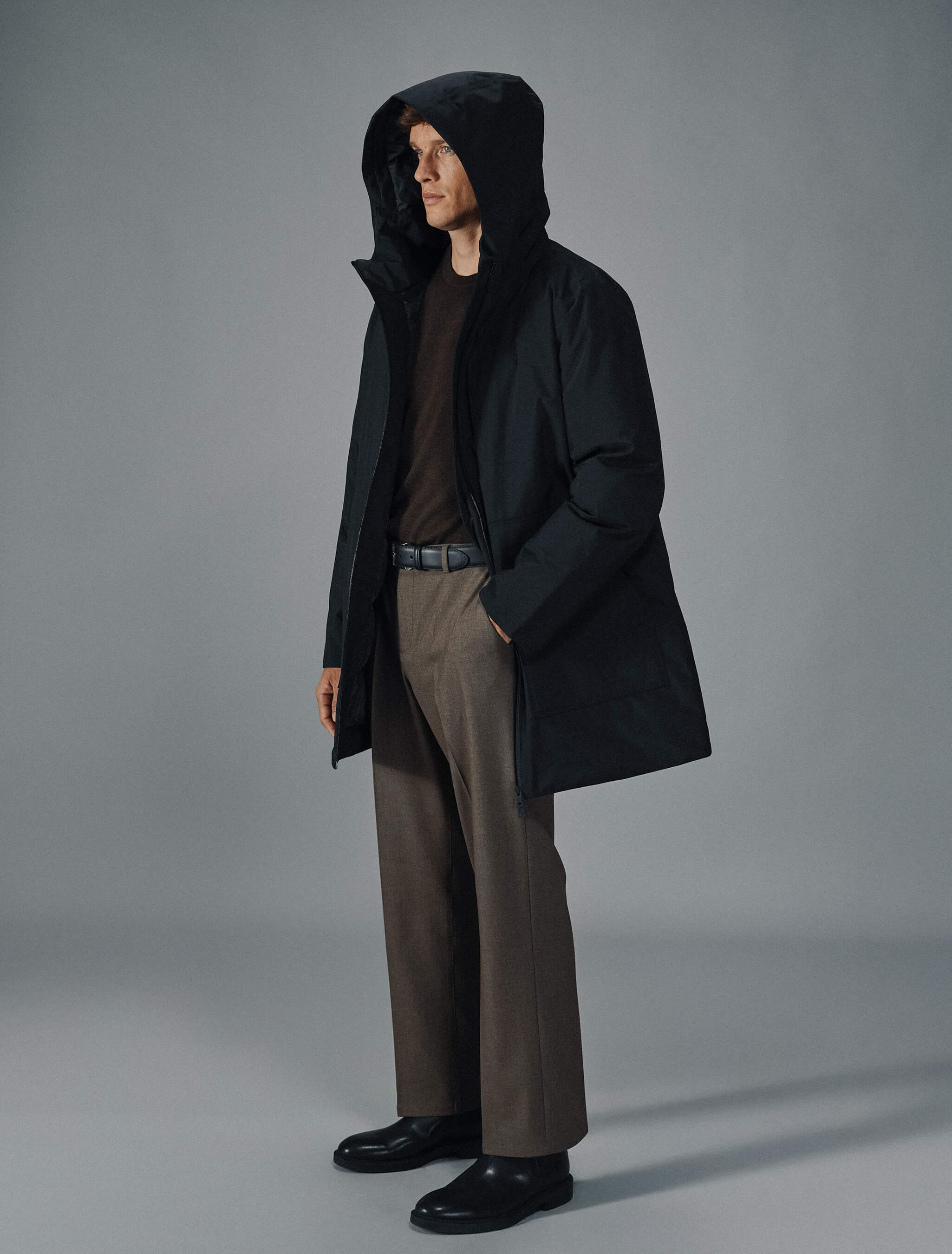 A man wears a black Utility Jacket Black over a dark brown sweater, brown trousers, and black shoes.